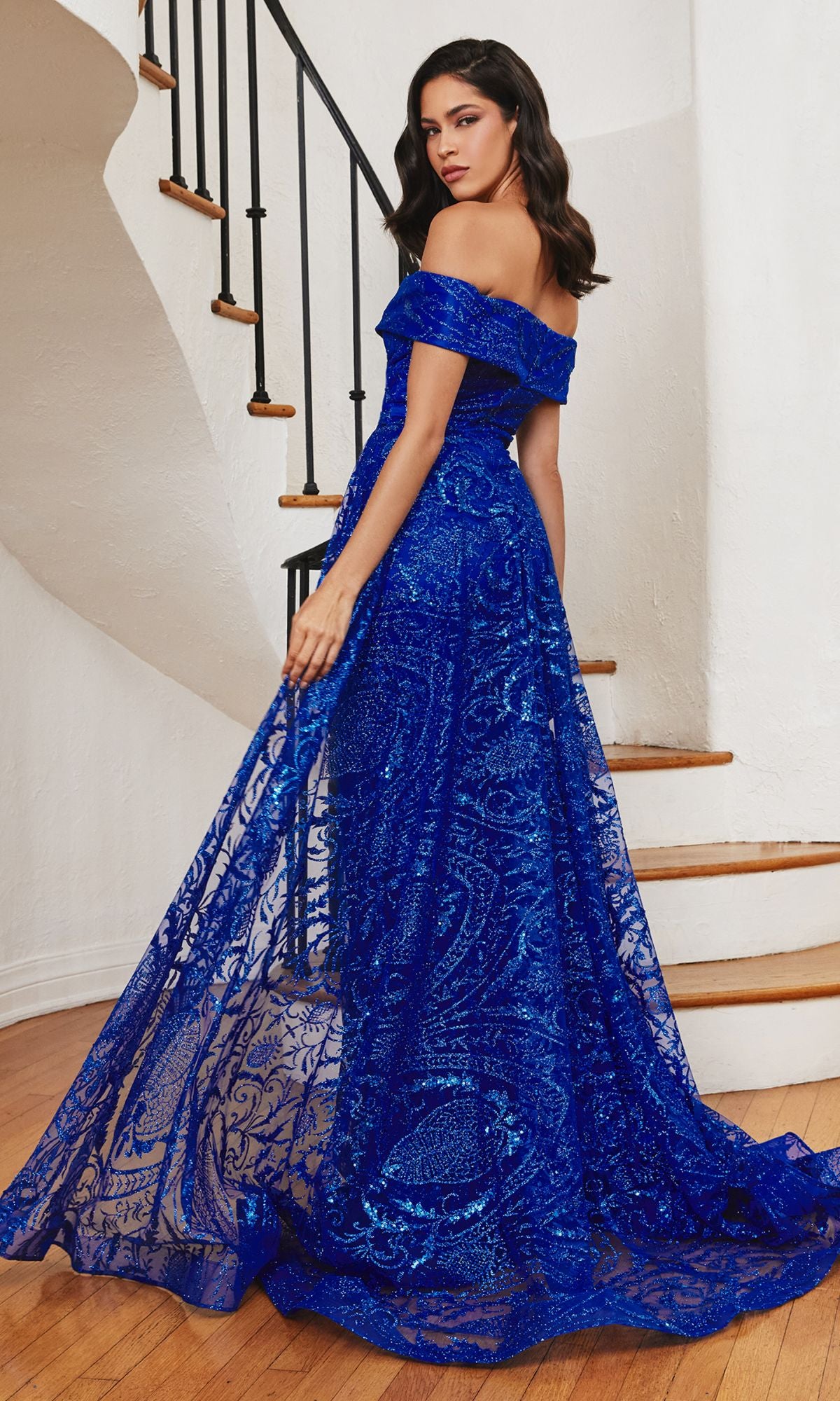 A woman stands on a staircase wearing the Long Prom Dress with Glitter-Print Overskirt J836, a floor-length royal blue gown featuring an off-the-shoulder design and elegant glitter-print overskirt.